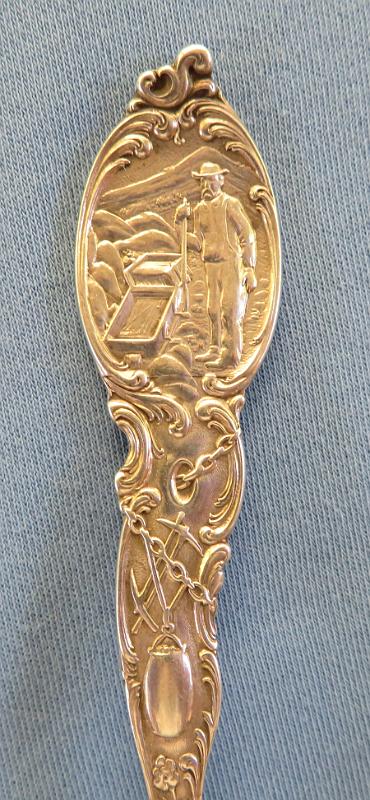Souvenir Mining Spoon Prescott AZ Handle.JPG - SOUVENIR MINING SPOON PRESCOTT AZ - Sterling silver souvenir spoon, features handle with miner and gold rocker box at top above picks and ore bucket, bowl embossed in deep relief with miner single jacking with artist marking H. OTTO in small letters near tip, back of handle marked PRESCOTT,ARIZONA at top above figure of cowboy on horse, back marked STERLING with makers mark of George E. Homer (Boston, MA 1875 - ?) and COPYRIGHTED, measures 5 1/2 in. length , weighs 37.3 gr. [Prescott is located in the Bradshaw Mountains of central Arizona, approximately 100 miles north of Phoenix.  The new town of Prescott was named in honor of historian William H. Prescott during a public meeting in 1864 and was officially incorporated in 1881. Prescott twice served as the capital of the Arizona Territory, once till November 1867 and again from 1877 to 1889 when the capital was moved to Phoenix.   Gold was discovered in creeks in the Agua Fria River Basin near Prescott in 1863.  Joseph Walker and a party of prospectors had set out from the California gold fields, possibly fleeing from conscription in the Confederate or the Union Army during the Civil War.  They found gold placers and, shortly afterward, lode deposits.  These discoveries were in what collectively became the Big Bug Mining District a few miles southeast of Prescott.  When the railroad arrived around 1898 transportation of equipment, minerals, and people became easier and cheaper.  Gold, silver, lead, and/or copper were produced from several famous mines including the Poland, McCabe, Silver Belt, Little Jessie, Henrietta/Big Bug, Blue Bell, Boggs/Iron Queen, and Iron King.  Several smelters were also built in the area.  The World War I era saw the greatest production from the district; however, the post-war drop in metal prices caused many mines to close.  The Iron King was the last of the major mines in the district.  After a series of different owners, it was operated by the Shattuck-Denn Mining Company from 1942 until it closed in 1969.  Prospectors staked claims in the Jerome District approximately 35 miles northeast of Prescott in 1876.  In 1882 the United Verde Copper Company was formed and mining started.  The rich oxidized ores produced copper, gold, and silver.  Transportation costs were very high until William A. Clark of Butte, Montana fame bought the company and brought the railroad to Jerome.  The United Verde Mine prospered and became the largest copper mine in the territory.  The original smelter built on unstable ground adjacent to the mine was replaced by a larger, more efficient one in Clarkdale.   In 1912, the Little Daisy Mine near the United Verde was purchased by James S. Douglas.  In 1914 and 1916 rich ore bodies were discovered.  A smelter was built in Clemenceau near the current town of Cottonwood.  The United Verde Extension Mining Company mined out the extension in 1930.  Phelps-Dodge purchased the United Verde and operated an open pit until mining ceased in 1953.  Today, the Douglas Mansion adjacent to the Little Daisy Shaft is part of the Jerome State Historic Park.  Jerome was designated a National Historic District in 1967.  Many of the historic buildings in Jerome have been converted to shops and eateries catering to tourists.  The Jerome Historical Society Museum occupies one of the buildings.  The Bagdad Mining District is located about 40 miles west of Prescott.  It has a rich history of mining including such famous mines as the Old Dick, Copper King, Copper Queen, and the Hillside among others.  The Bagdad deposit was discovered in 1862 and the claims were patented in 1889.  It was owned by a series of companies.  Exploration drilling was started in 1919 and the first mill was constructed in 1928.  During World War II, a 2000 ton per day mill was constructed with funds from the Reconstruction Finance Corporation. In the late 1940’s, the underground mine using block caving was converted to an open pit.  The town of Bagdad was a company town.  In 1973 the Bagdad Copper Corp. merged with the Cyprus Mines Corp. to form the Cyprus Bagdad Copper Company.  It later became part of Phelps-Dodge.  As a result of the 2007 merger, the mine is now operated by Freeport-McMoRan Copper and Gold Corporation. ] 